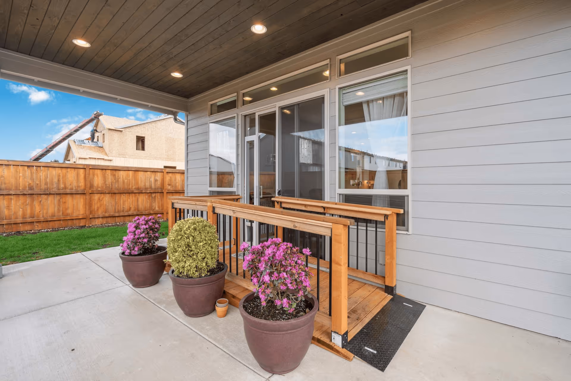 Covered patio with a wooden ramp and potted flowering plants in front of sliding glass doors.