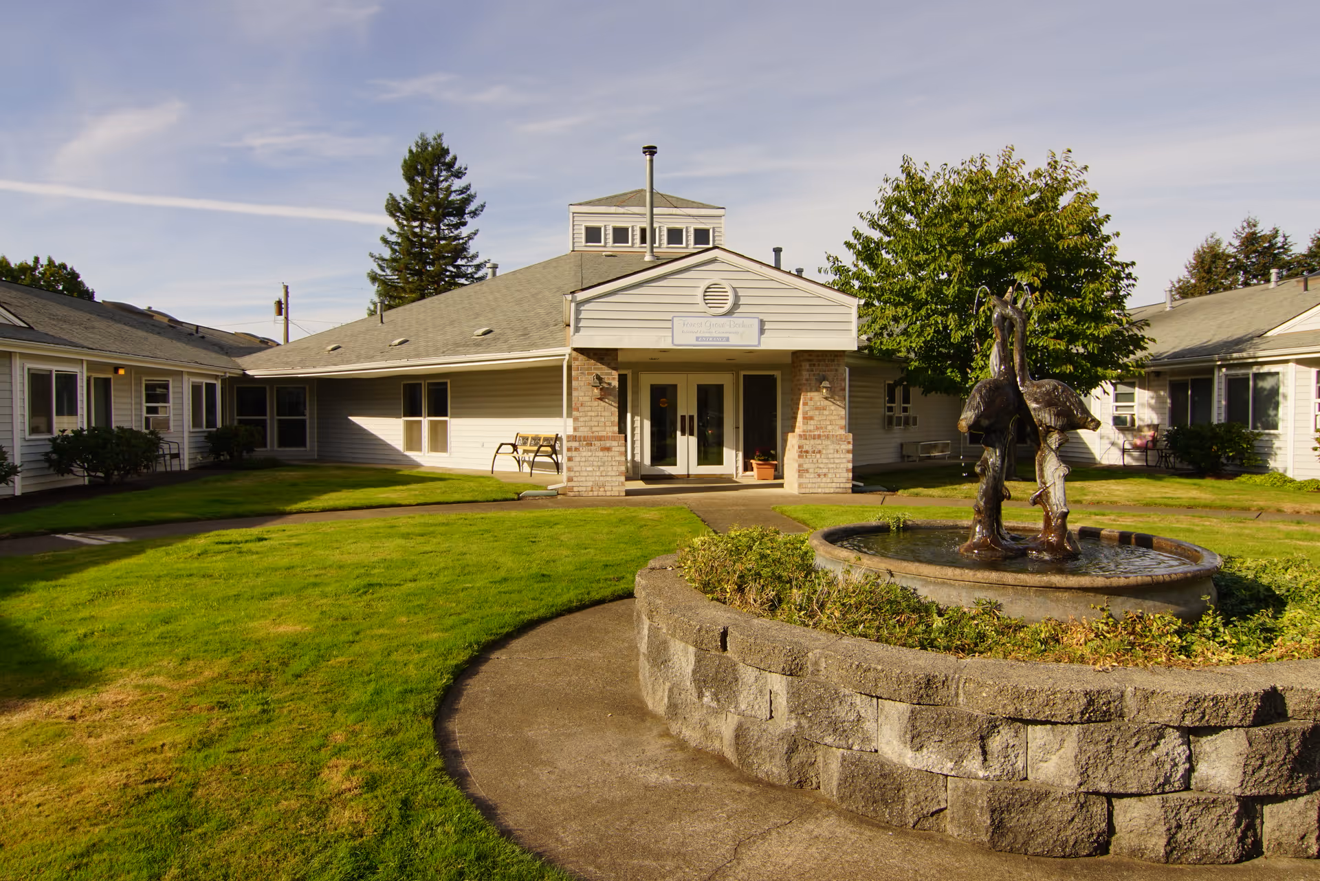 Exterior view of Forest Grove Beehive Assisted Living facility showing a single-story building with a central entrance, surrounded by green lawns and a circular stone planter with a bird sculpture fountain in the foreground.