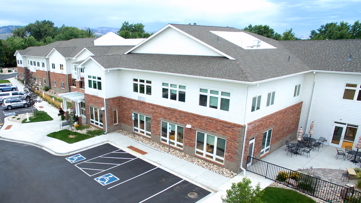 Exterior view of New Mercer Commons Assisted Living facility showing a two-story building with white siding and red brick accents. The building has multiple windows and a gray shingled roof. There is a parking lot with designated handicapped parking spaces in front, and an outdoor patio area with tables and chairs on the right side. Trees and greenery surround the property with mountains visible in the background under a partly cloudy sky.