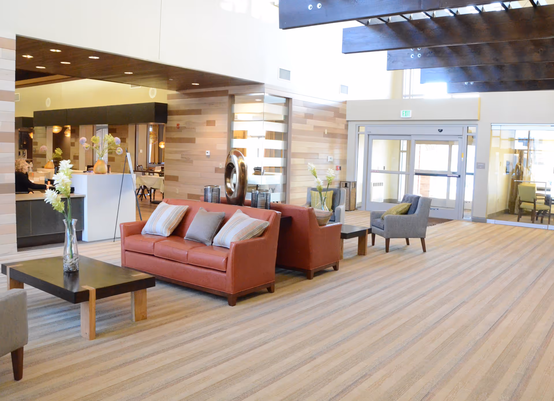 A spacious and well-lit senior living facility common area with modern furniture including a red sofa with pillows, a coffee table with a vase of flowers, and gray armchairs. The room features wood paneling on the walls, large windows, and a high ceiling with exposed beams. In the background, there is a reception desk and a dining area.