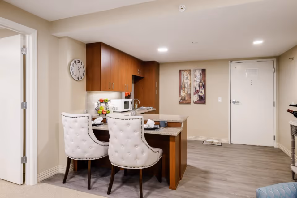 Interior view of a small kitchen area with wooden cabinets, a granite countertop with two white cushioned chairs, a microwave, a fruit basket, and a wall clock. The room has light-colored walls, two paintings on the wall, a white door, and wood flooring.