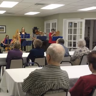 A group of elderly people seated at tables watching a small ensemble of musicians playing string instruments in a room with light green walls and white ceiling tiles.