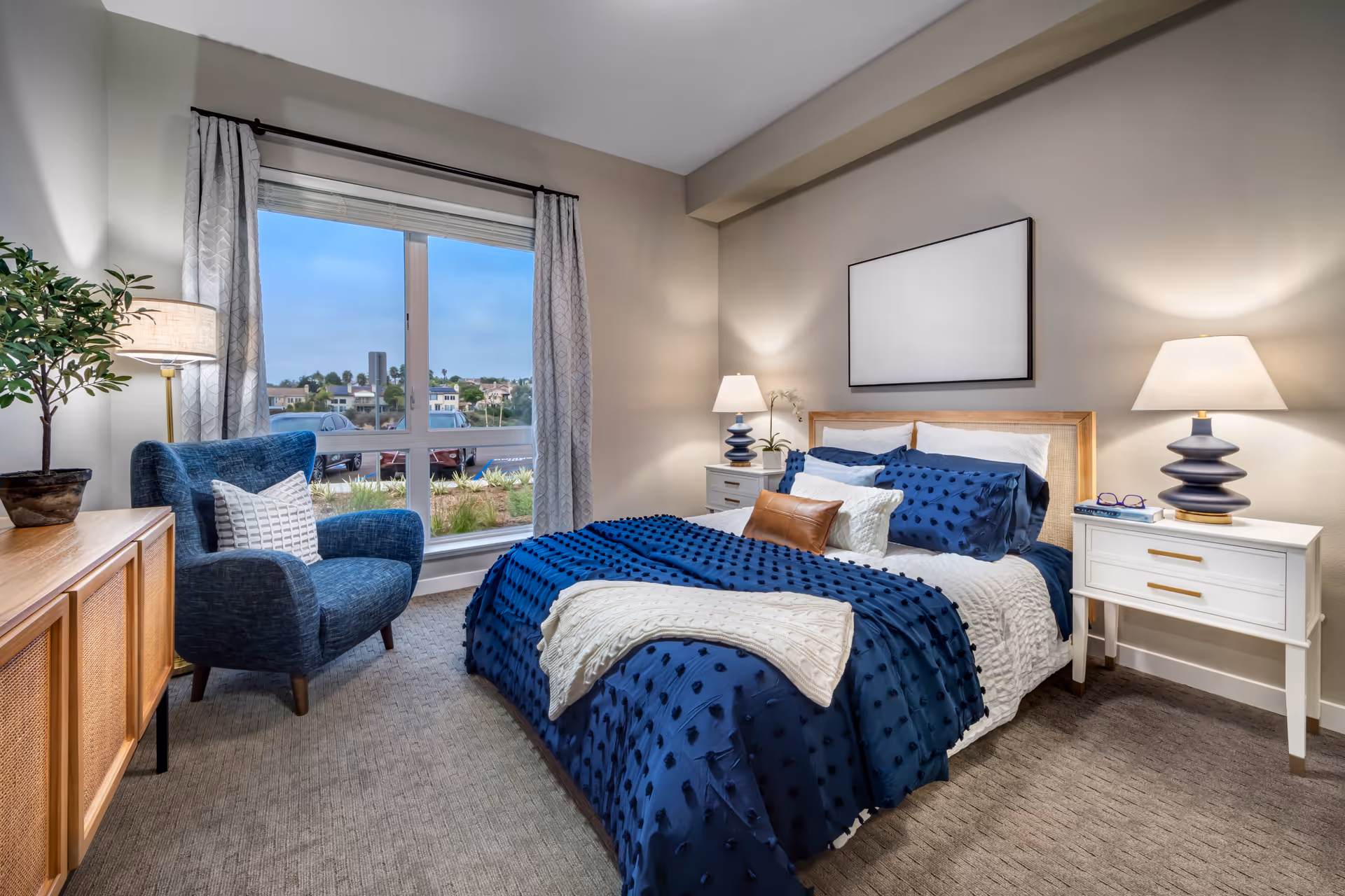 A cozy bedroom with a large window showing an outdoor view. The room features a bed with blue and white bedding, two white nightstands with matching lamps, a blue armchair with a white pillow, a wooden dresser with a potted plant, and neutral-colored walls and carpet.