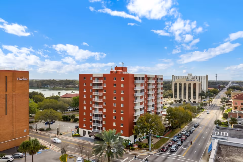 Red mid-rise apartment building with balconies on a city street near a lake under a blue sky.