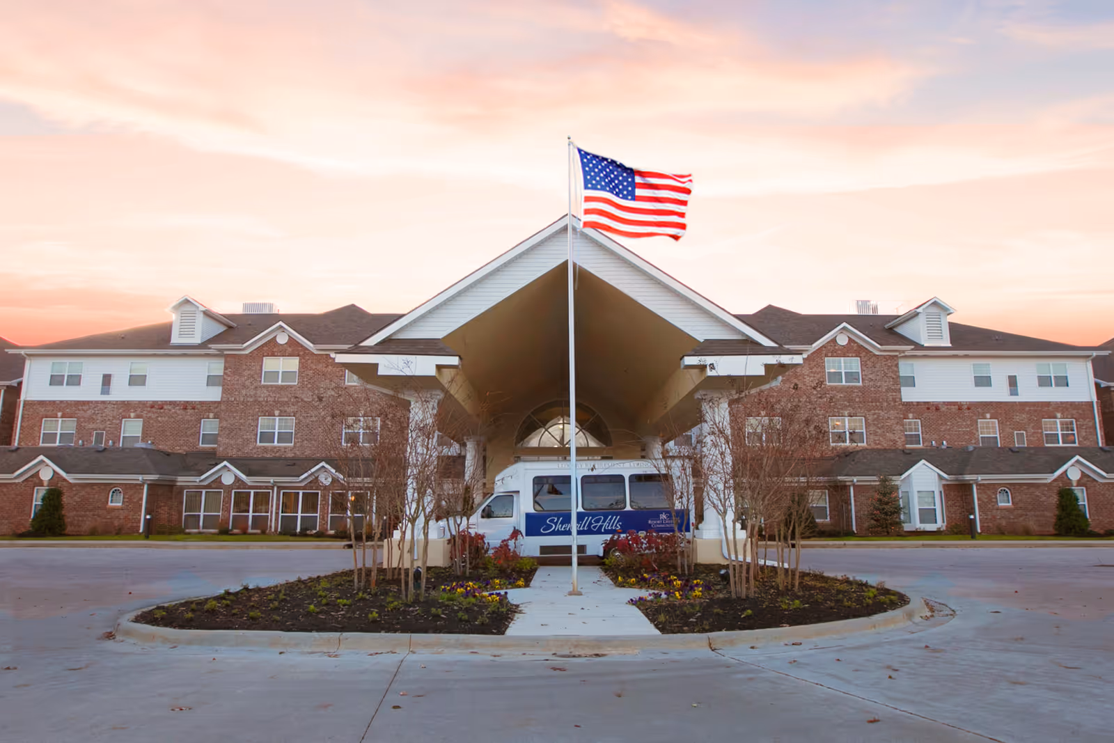 Front exterior view of Sherrill Hills Retirement Resort building at sunset with an American flag on a flagpole in the center and a shuttle van parked under the entrance canopy.