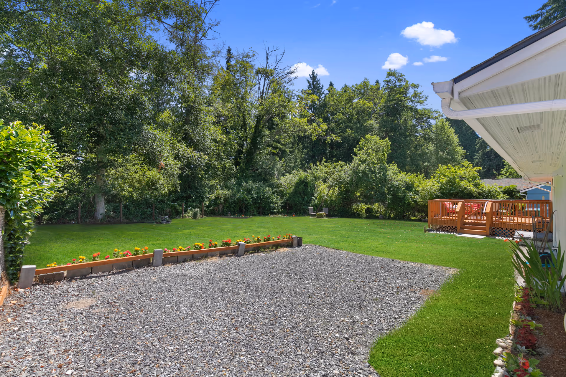 A spacious outdoor area with a gravel section in the foreground, bordered by a small flower bed with colorful flowers. There is a well-maintained green lawn extending towards a dense line of trees and shrubs in the background. On the right side, part of a building with a white roof overhang and a wooden deck with outdoor seating is visible under a bright blue sky with a few clouds.