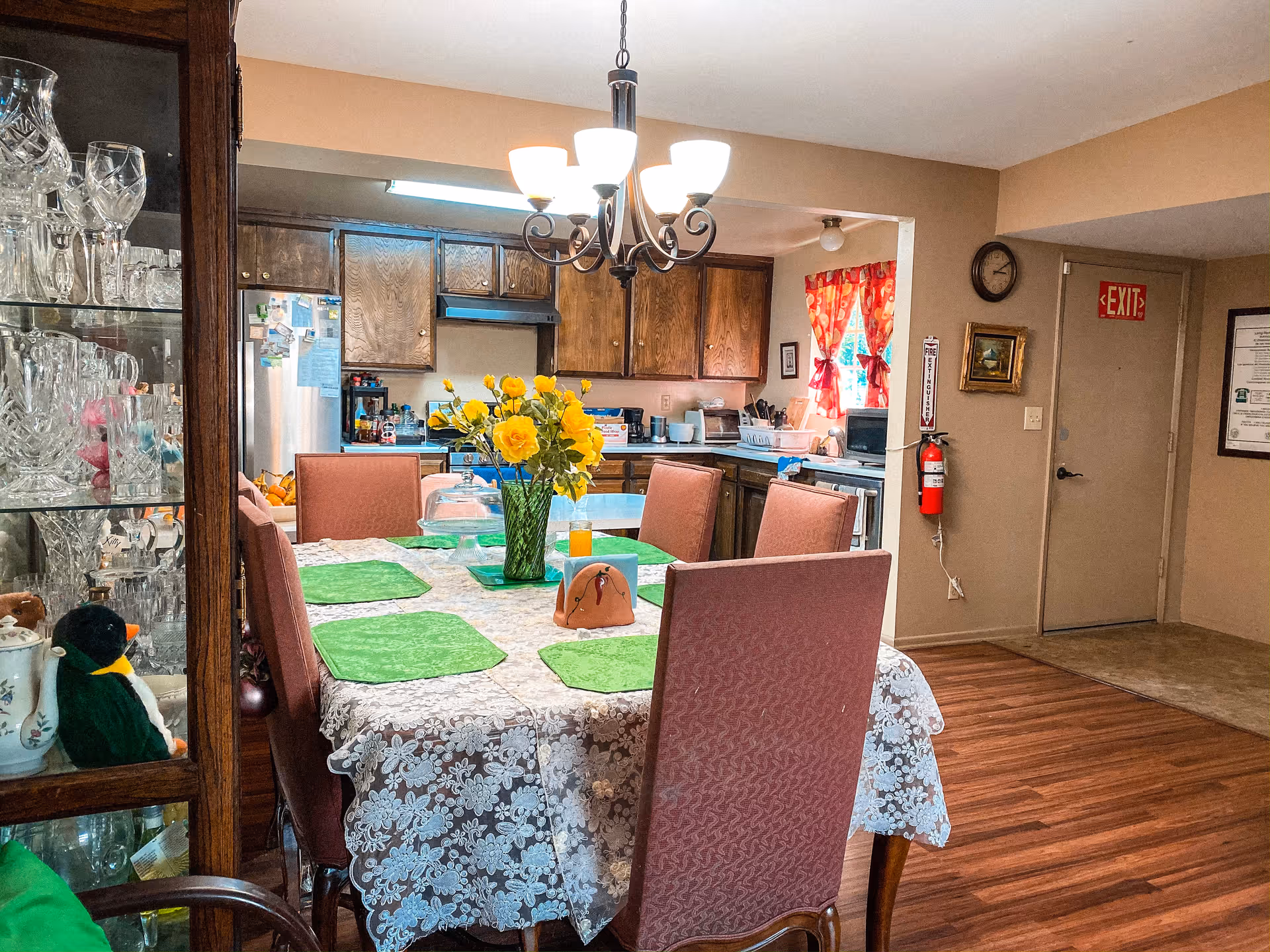 Dining room and kitchen interior with a table set with green placemats and a vase of yellow flowers, surrounding chairs, chandelier, and a glassware cabinet.