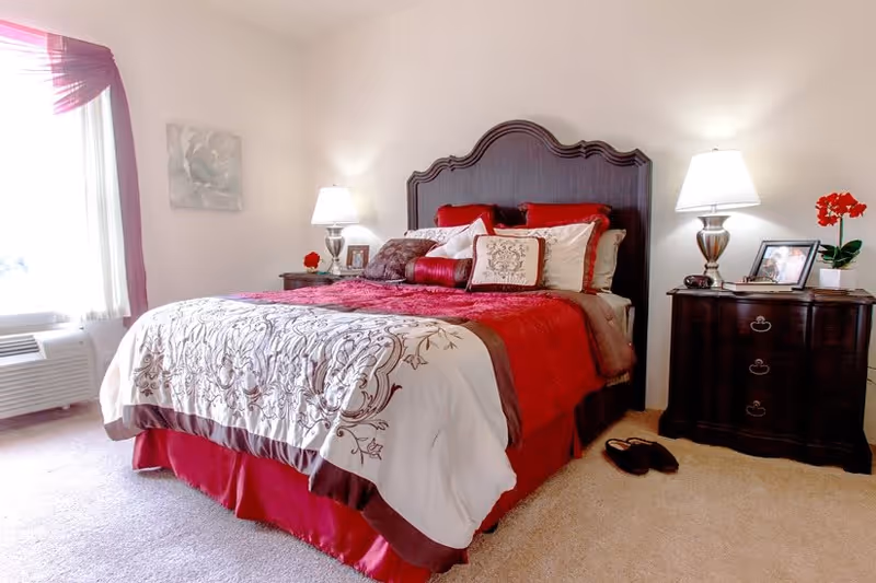 A neatly made bed with a dark wooden headboard in a bedroom. The bed has a red and white patterned comforter with multiple pillows. On either side of the bed are dark wooden nightstands with lamps, framed photos, and a red flower. There is a window with sheer curtains and a red valance on the left side, and a pair of slippers on the carpeted floor near the right nightstand.