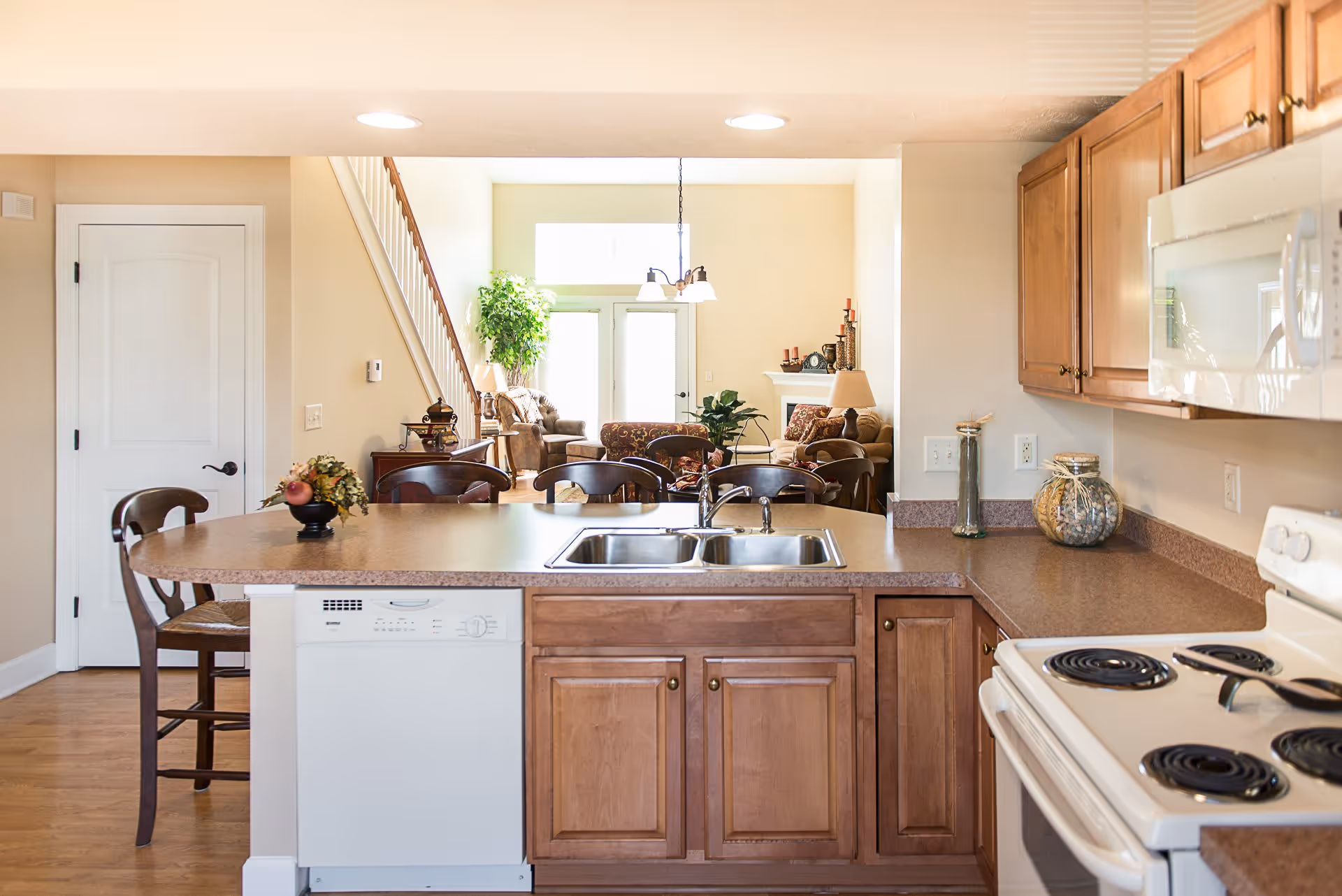 View of a kitchen with wooden cabinets, a white electric stove, a white dishwasher, and a double sink on a countertop. Beyond the kitchen is a living room area with chairs, a sofa, a fireplace, plants, and a staircase to the left.
