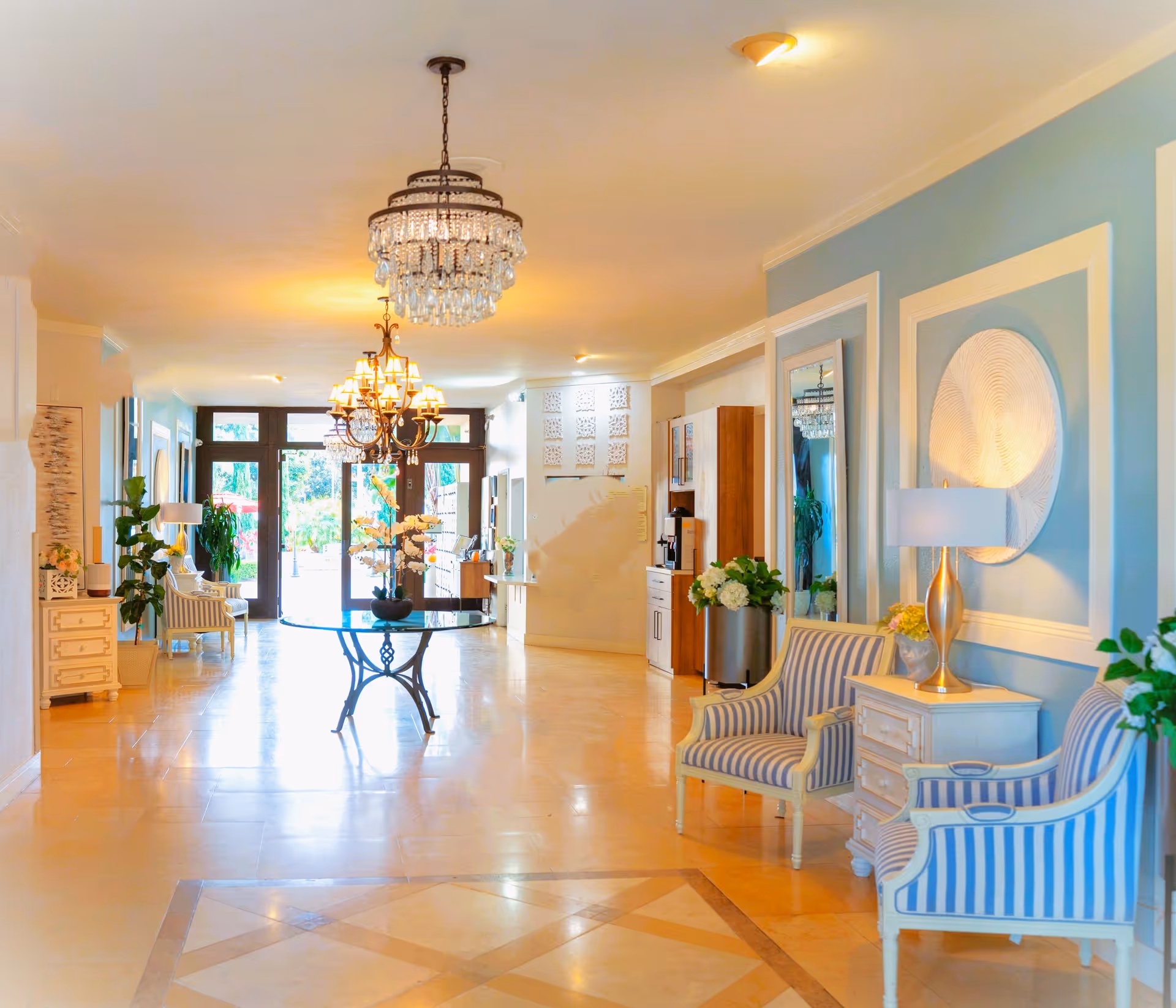 Bright, elegant retirement community lobby with chandeliers, striped armchairs, console tables, and a central round table.