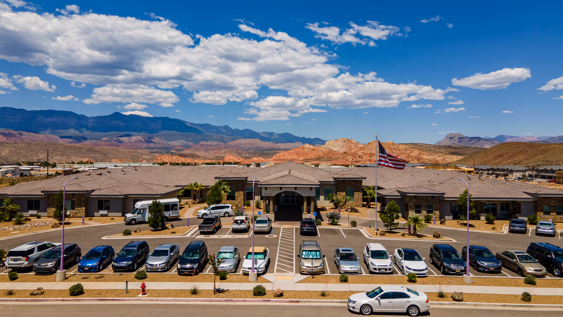 Front view of a single-story senior living facility with an American flag and parked cars in front, set against red rock mountains and a blue sky.