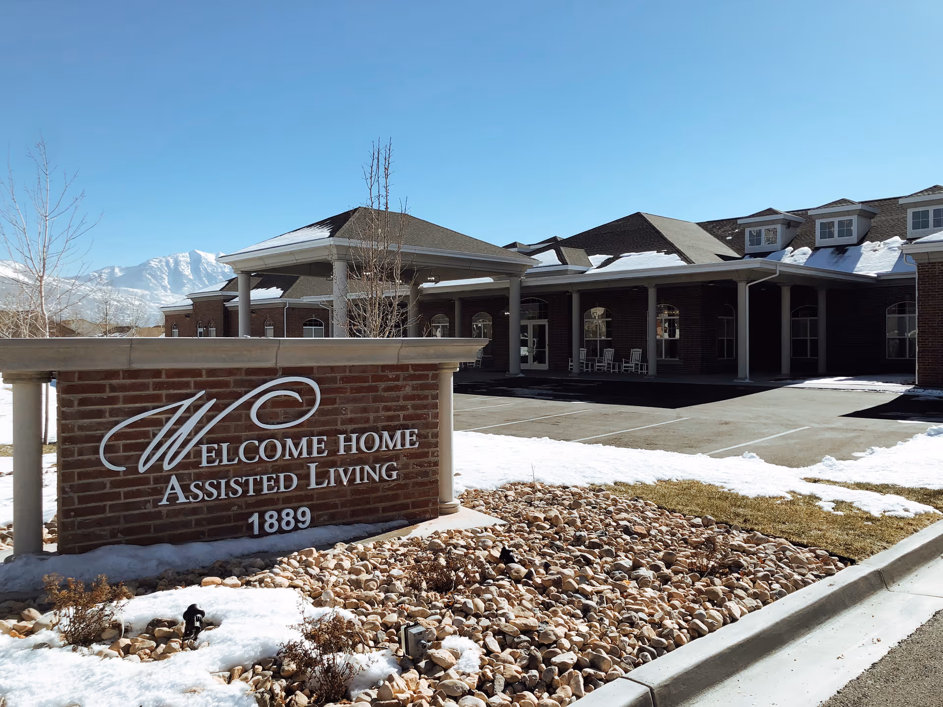 Brick sign reading "Welcome Home Assisted Living 1889" in front of the facility's entrance and parking area with patches of snow.