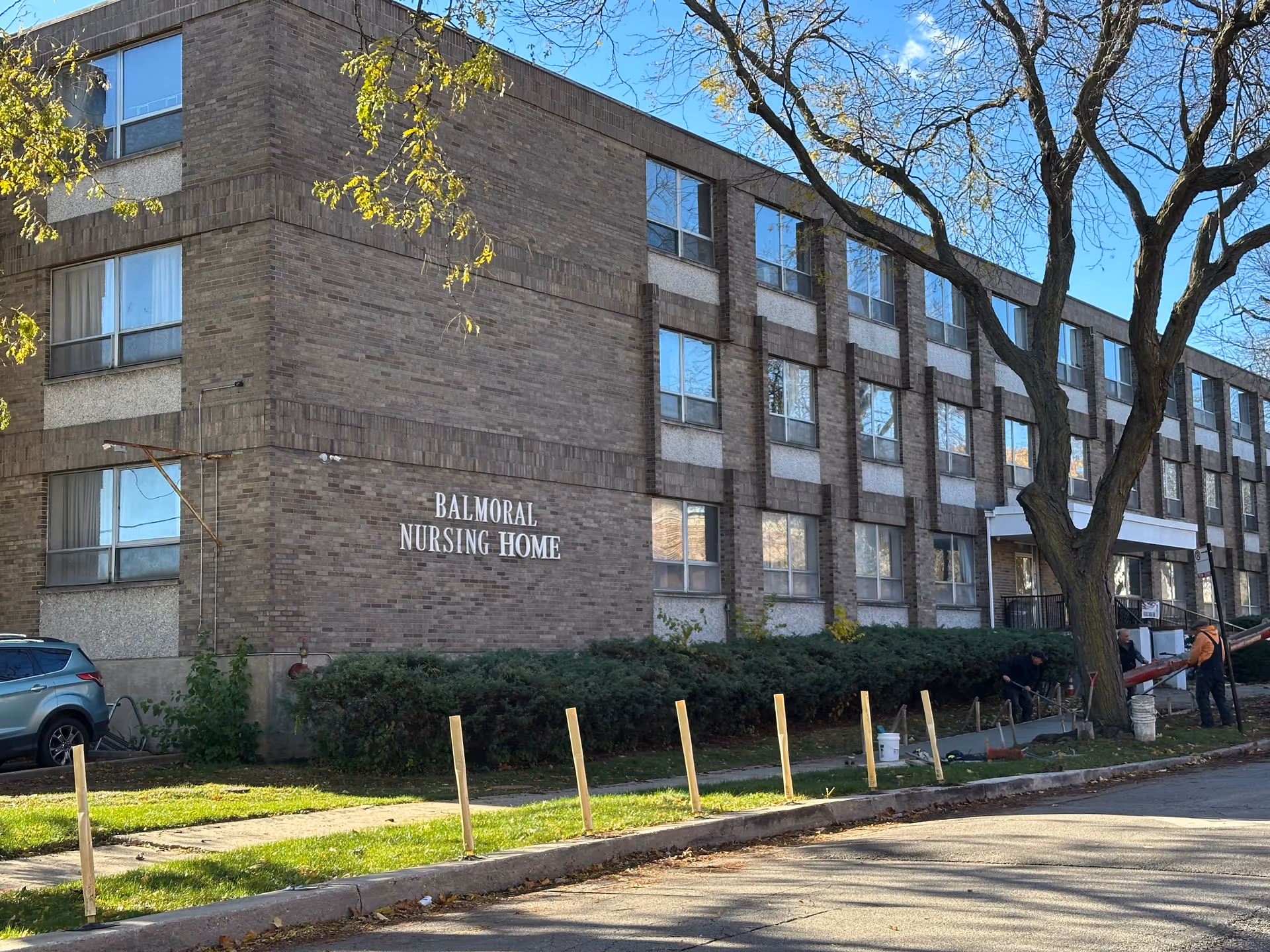 Exterior view of Balmoral Nursing Home, a three-story brick building with multiple windows. There are some bushes along the front and a large tree on the right side. Workers are seen doing construction or maintenance work on the sidewalk in front of the building. The sky is clear and blue.