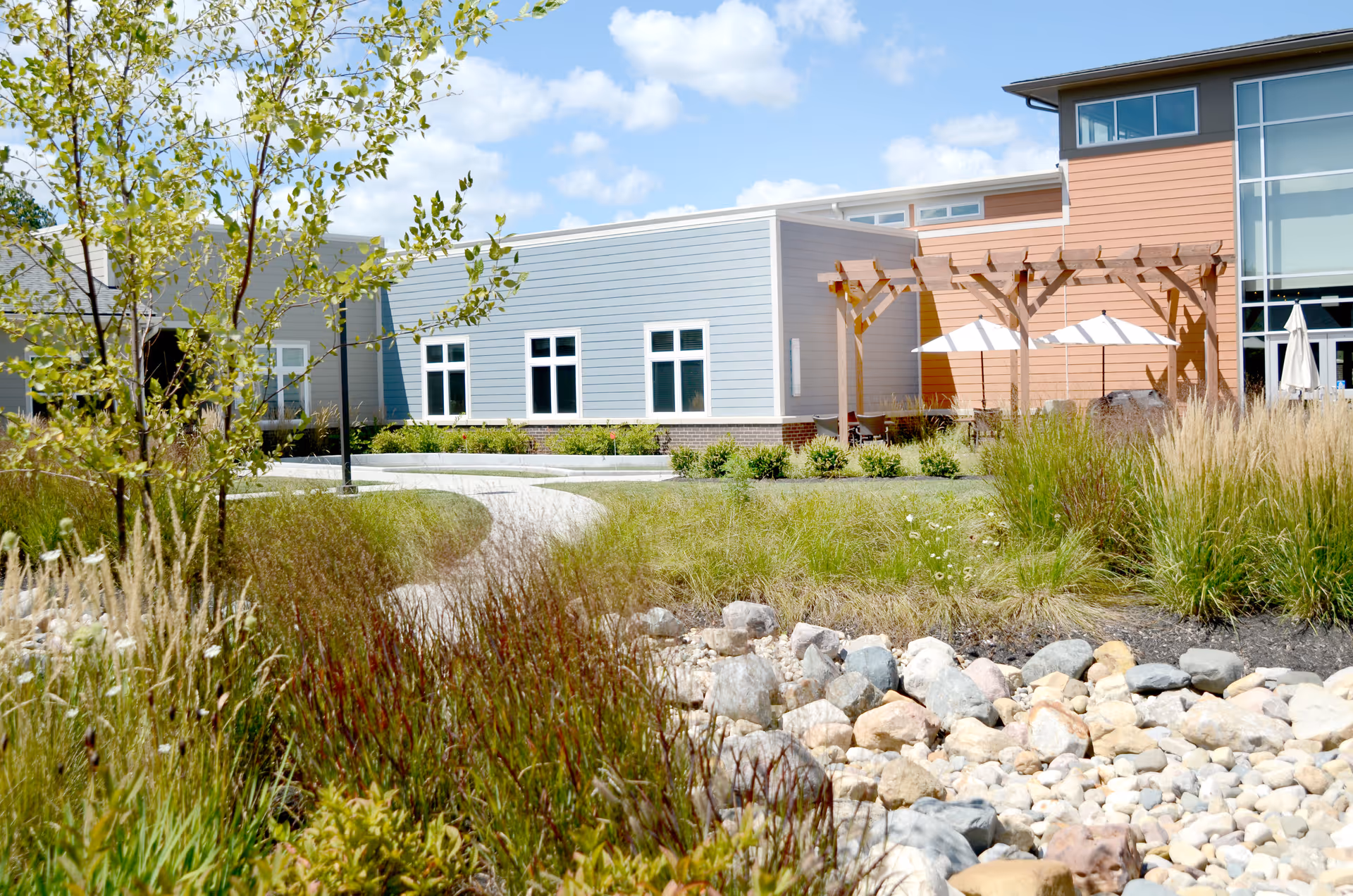Outdoor garden area at Wellbrooke of Avon featuring a winding pathway, various grasses and plants, a rock bed, and a building with blue and orange siding. There is a wooden pergola with white umbrellas providing shade over seating areas.