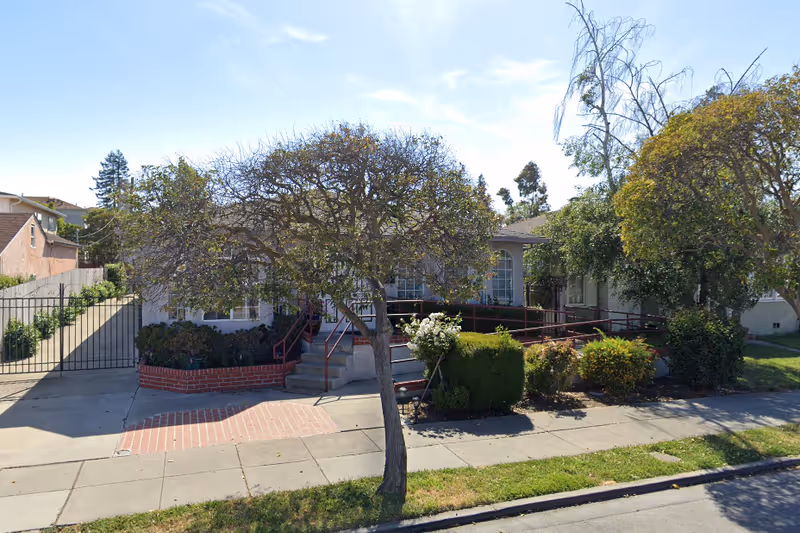 Exterior view of a single-story building with a ramp and stairs leading to the entrance, surrounded by trees and bushes, with a gated driveway on the left side.