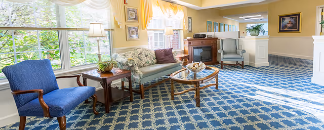 Sunlit sitting area with upholstered chairs, a floral sofa, glass-top coffee table and large windows overlooking greenery.