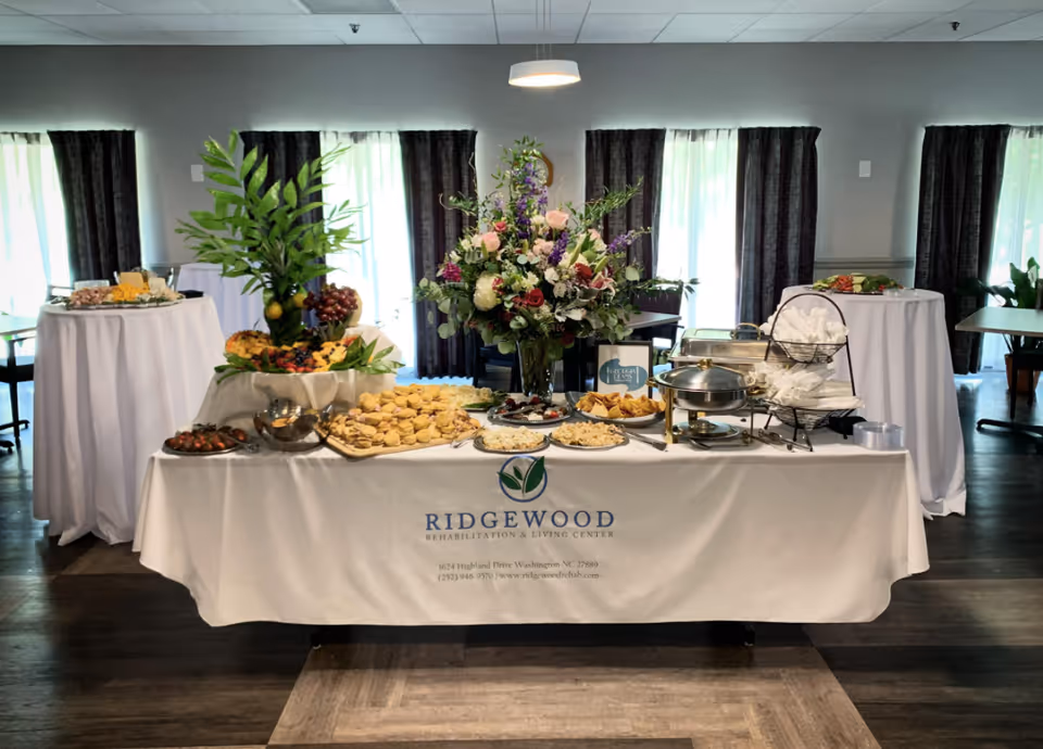 A buffet table set up with a variety of food including sandwiches, fruit, and appetizers, decorated with a large floral arrangement in the center. The table is covered with a white tablecloth featuring the Ridgewood Rehabilitation & Living Center logo and contact information. The room has large windows with dark curtains and additional round tables with white tablecloths in the background.