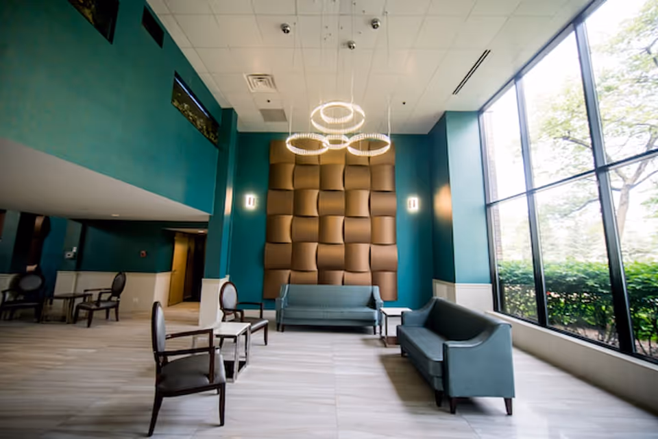 Spacious senior living lobby with teal walls, a textured wall panel, sofas and chairs by large floor-to-ceiling windows and modern circular chandeliers.