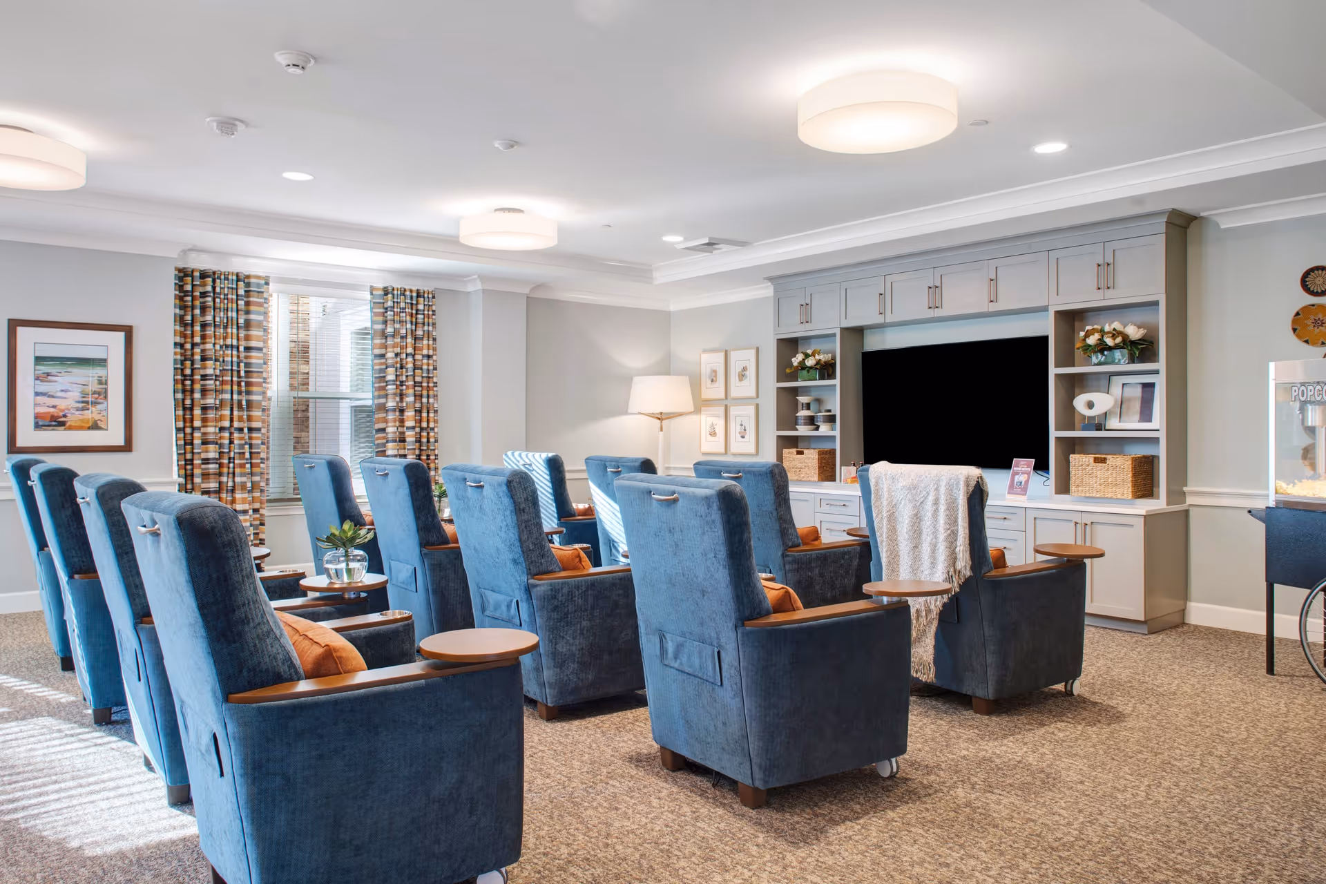 A cozy common area with two rows of blue upholstered armchairs facing a large flat-screen TV mounted on a built-in gray entertainment center. The room has patterned curtains on the windows, a floor lamp, framed artwork on the walls, and a popcorn machine on the right side. The space is well-lit with ceiling lights and natural light from the windows.