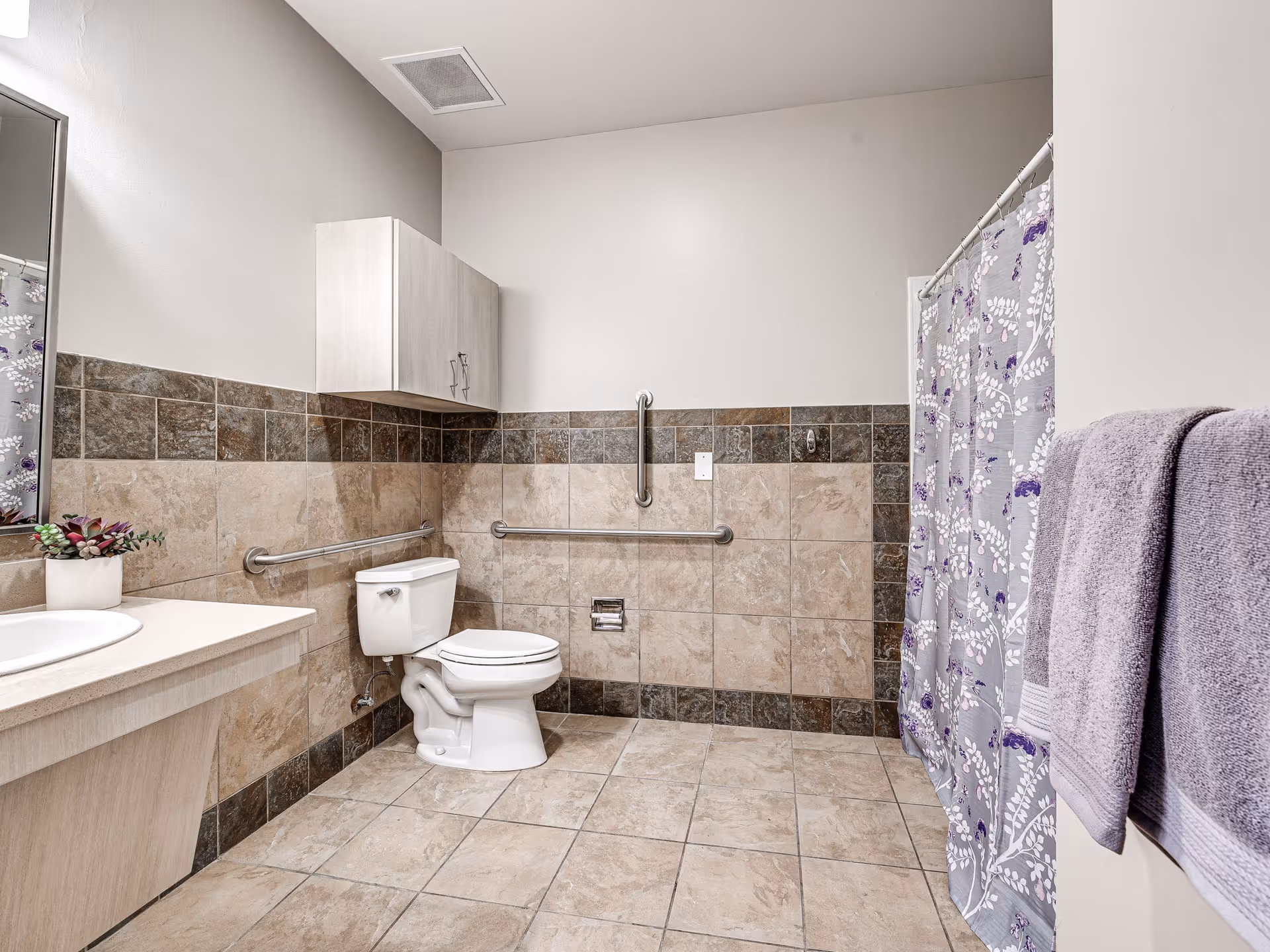 A clean, spacious bathroom with beige and brown tiled walls and floor. The bathroom features a white toilet with grab bars on the walls for accessibility, a sink with a countertop and a small potted plant, a wall-mounted cabinet above the toilet, a shower area with a floral-patterned shower curtain, and two folded purple towels hanging on a towel rack.