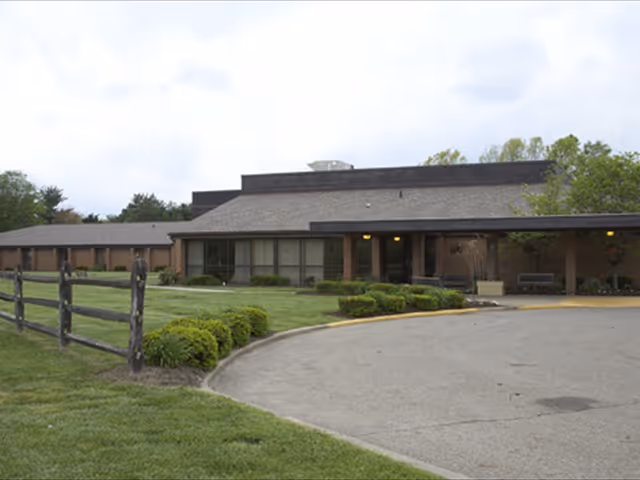 Exterior view of a single-story senior living facility building with a sloped roof, large windows, and a covered entrance. The building is surrounded by a well-maintained lawn, shrubs, and a wooden fence. The sky is cloudy.