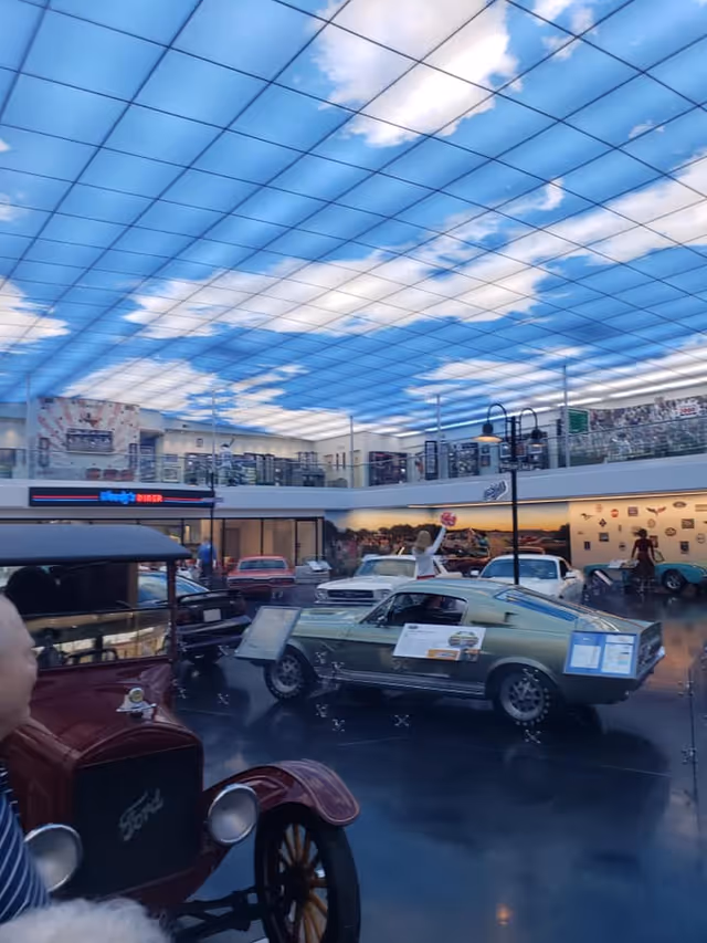 Indoor exhibit featuring vintage cars displayed on a shiny black floor under a ceiling designed to look like a blue sky with white clouds. The space has a second-floor balcony with railings and various decorations on the walls, including a sign for a diner. A person is partially visible in the foreground.