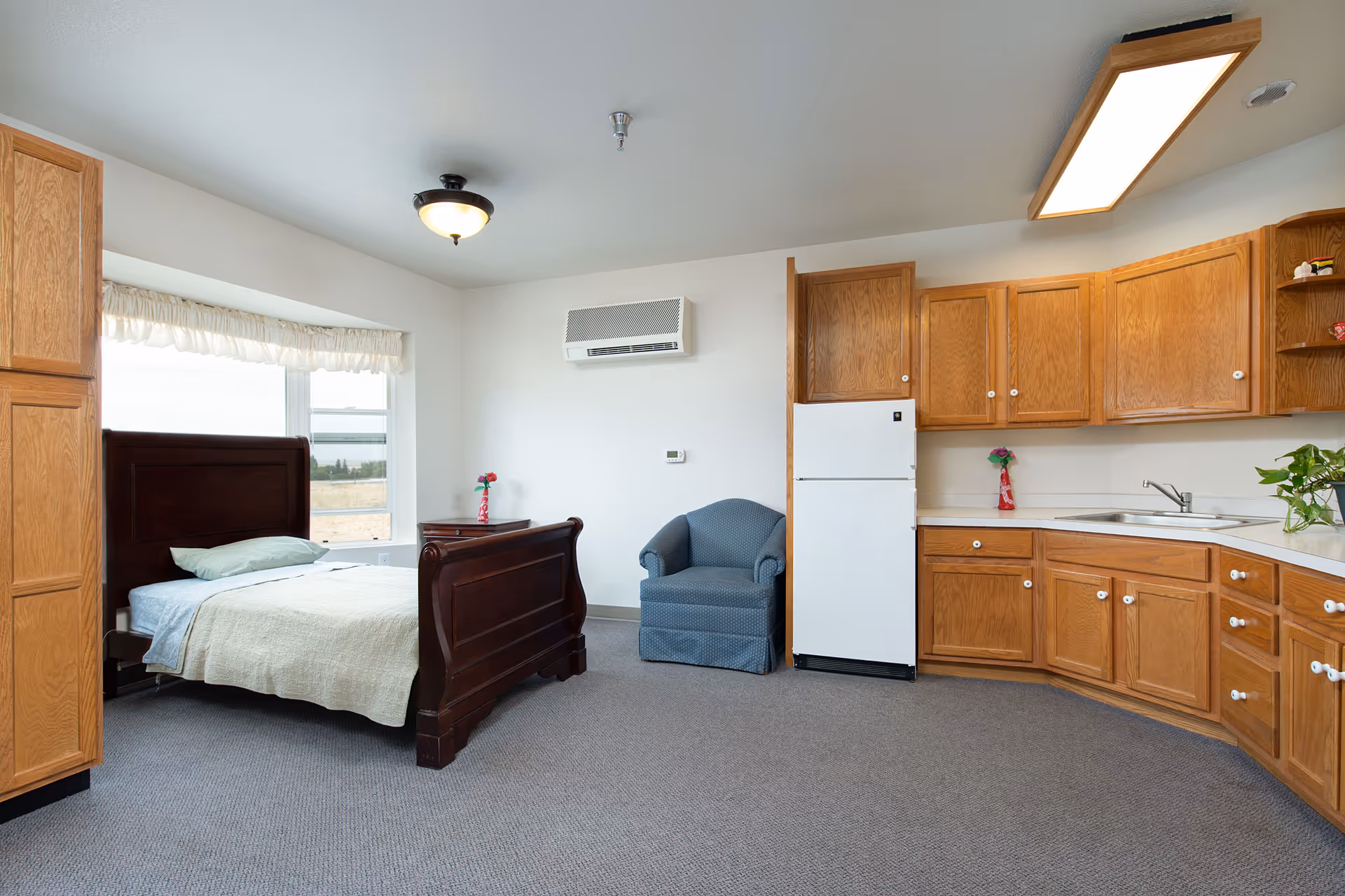 A senior living facility room with a wooden bed, a blue armchair, a small wooden nightstand with a flower vase, and a kitchenette with wooden cabinets, a white refrigerator, and a sink. The room has a large window with a white valance and carpeted floor.
