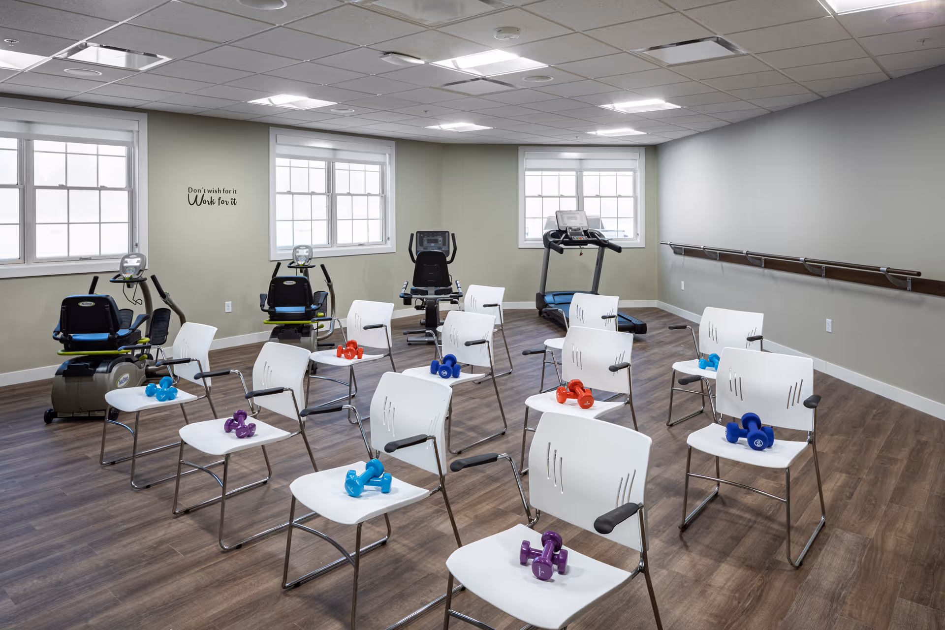 A fitness room with several white chairs arranged in rows, each holding pairs of colorful dumbbells. In the background, there are exercise machines including stationary bikes and a treadmill. The room has large windows, wood flooring, and a motivational quote on the wall that reads 'Don't wish for it Work for it'.