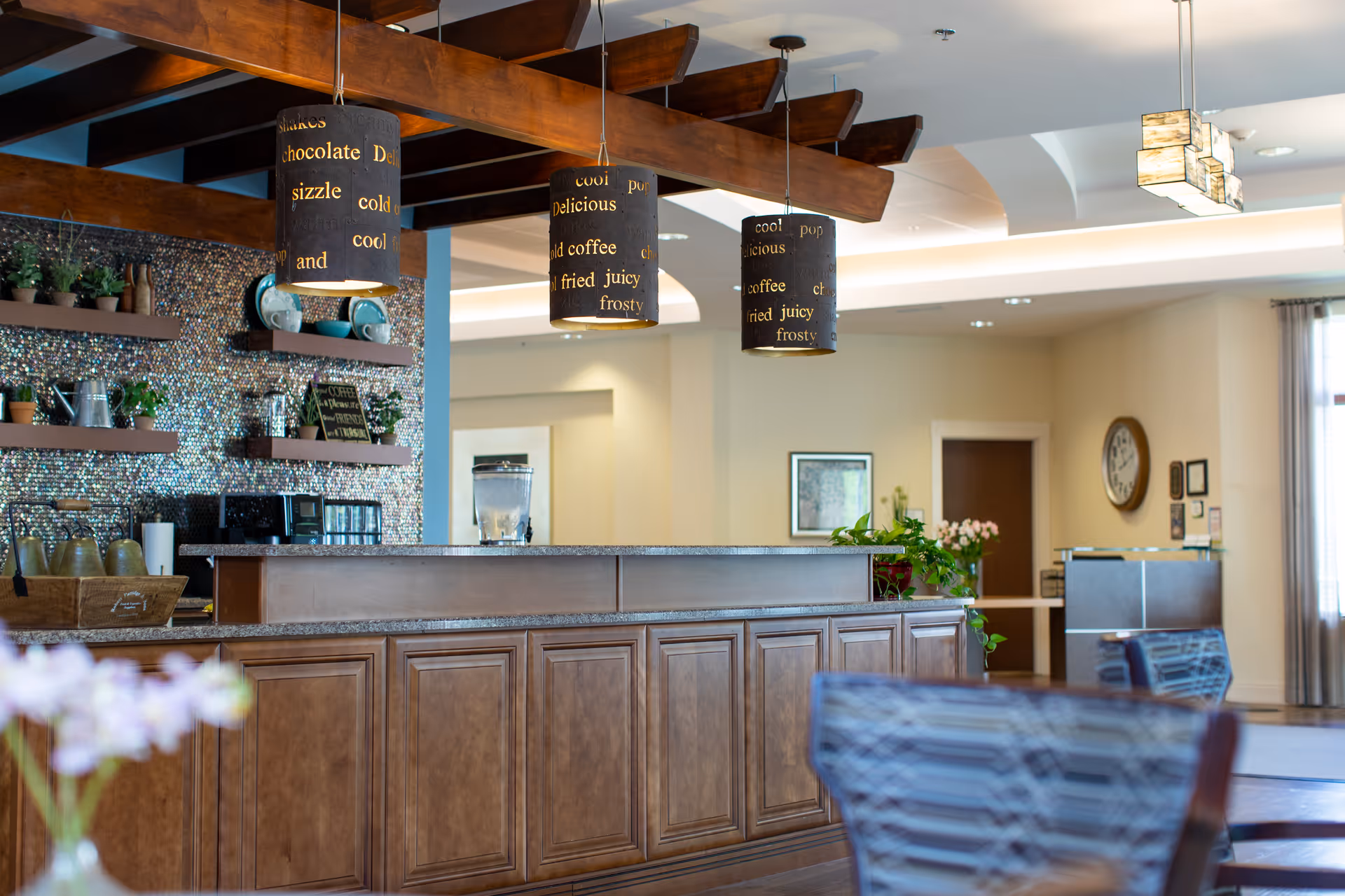 Interior view of a reception or common area with a wooden counter, three hanging pendant lights with words related to food and drinks, shelves with plants and decorative items, and seating in the foreground. The space has warm lighting and a welcoming atmosphere.
