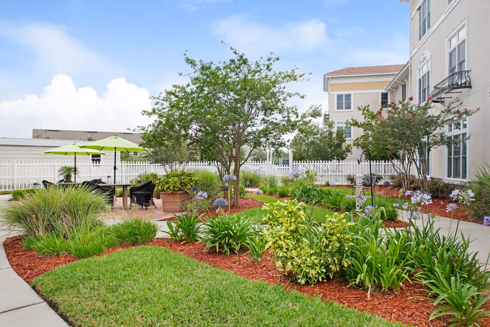 Well-maintained outdoor courtyard with landscaped flowerbeds, trees, a seating area with green umbrellas, and a white fence beside a multi-story building.