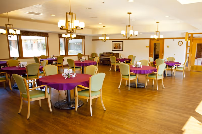 A bright dining room with multiple round tables covered with purple tablecloths, each surrounded by green cushioned chairs. The room has wooden flooring, large windows letting in natural light, modern ceiling lights, and a piano against the far wall.