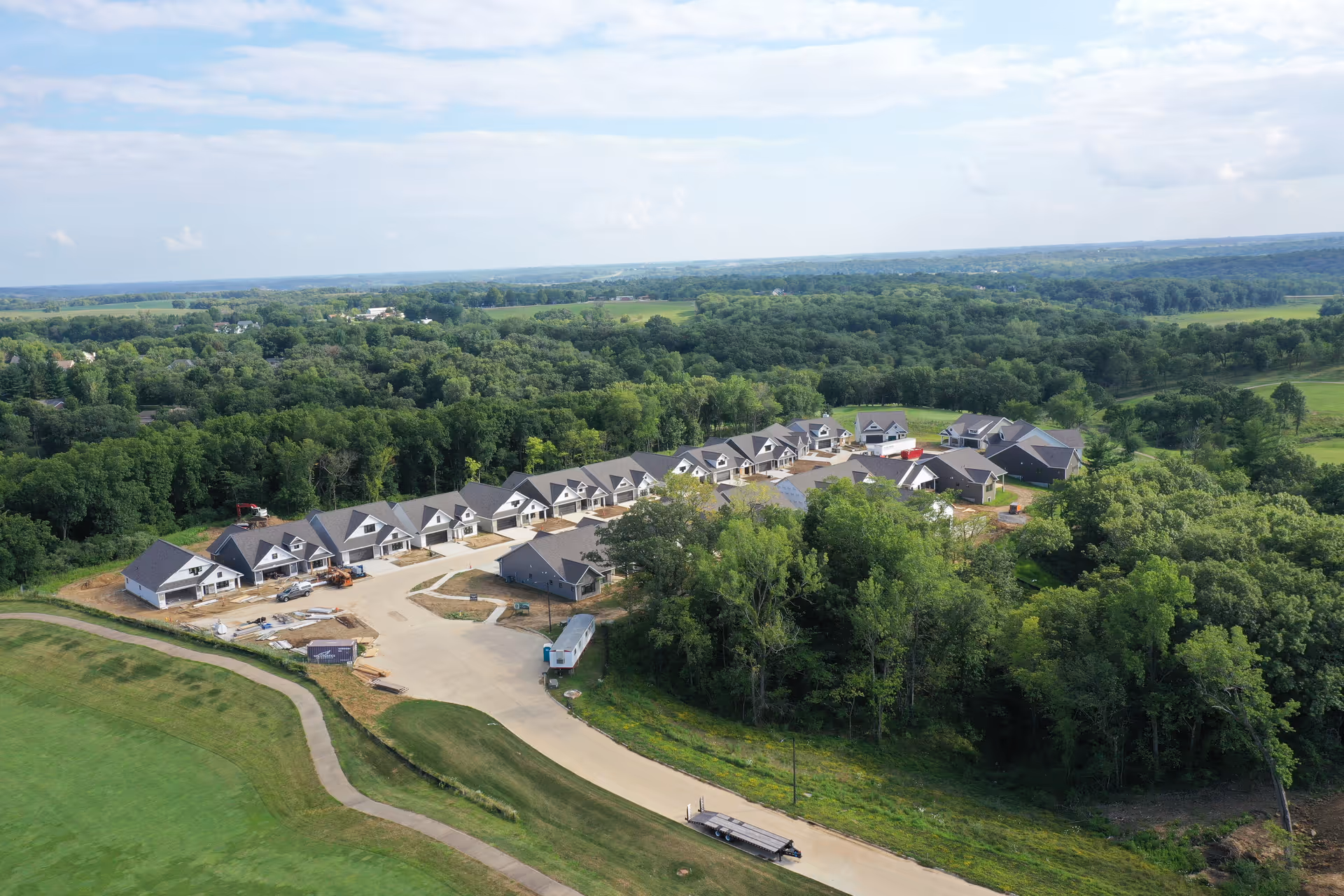 Aerial view of a residential community under construction surrounded by dense green trees and open fields, featuring multiple newly built houses with gray roofs arranged along a curved road.