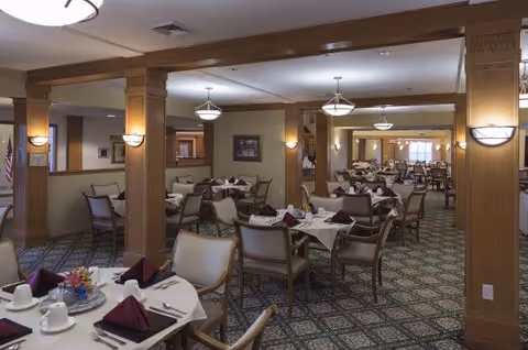 A spacious dining room in a senior living facility with multiple round tables covered with white tablecloths, each set with plates, cups, silverware, and folded burgundy napkins. The room features wooden columns, patterned carpet, and soft lighting from ceiling fixtures and wall sconces.