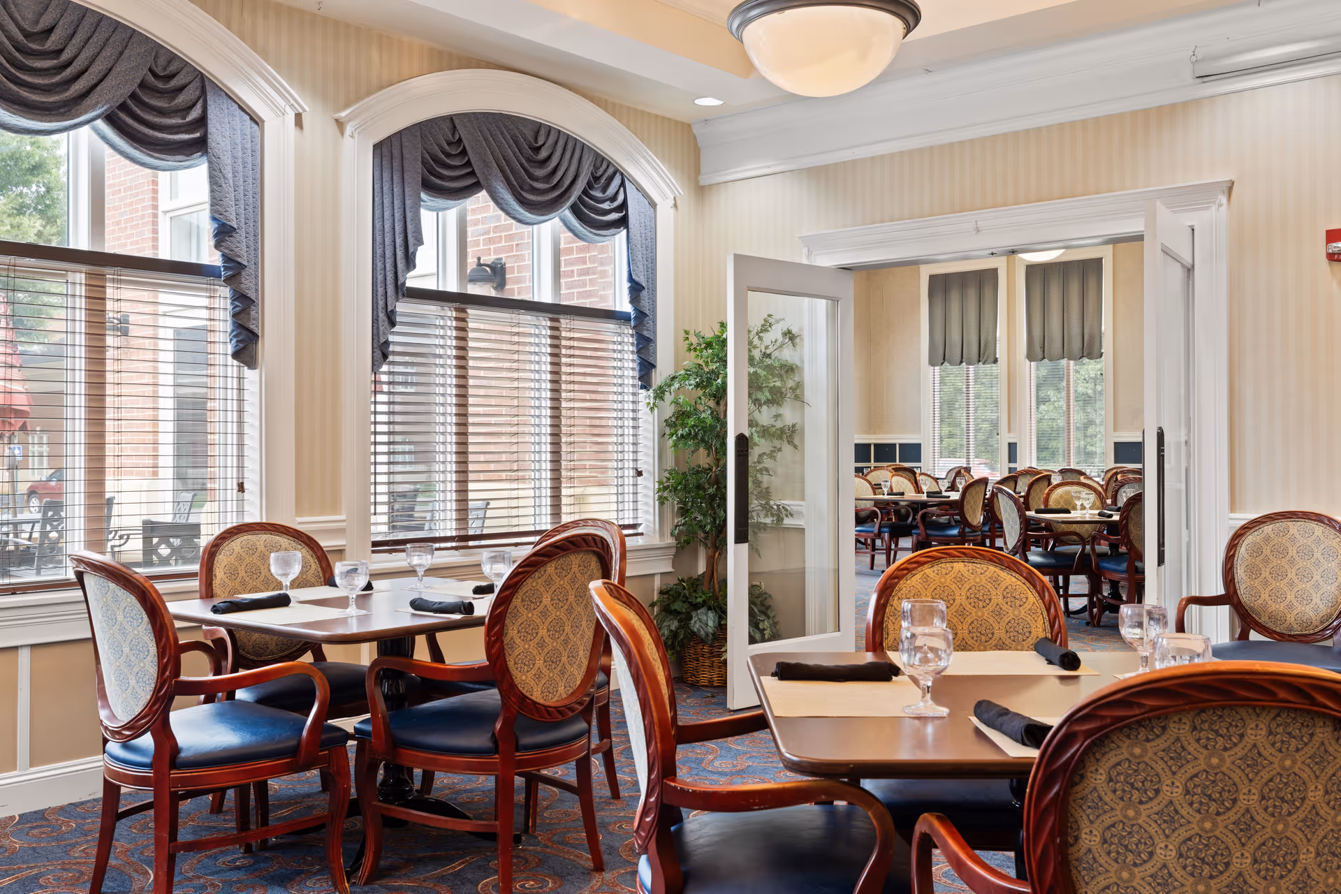 A dining room with wooden tables and upholstered chairs arranged neatly. Each table is set with placemats, folded black napkins, and water glasses. Large windows with blue drapes and blinds allow natural light to fill the room. A glass door leads to another dining area with similar furniture and window treatments. A potted plant is placed near the door.