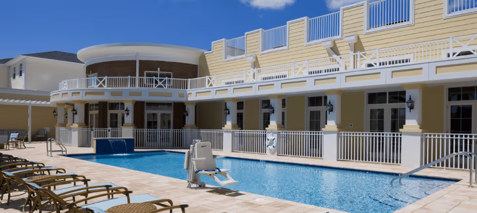 Outdoor swimming pool area at Watercrest Buena Vista with several lounge chairs lined up along the poolside, a pool lift for accessibility, and a two-story building with balconies and railings in the background under a clear blue sky.
