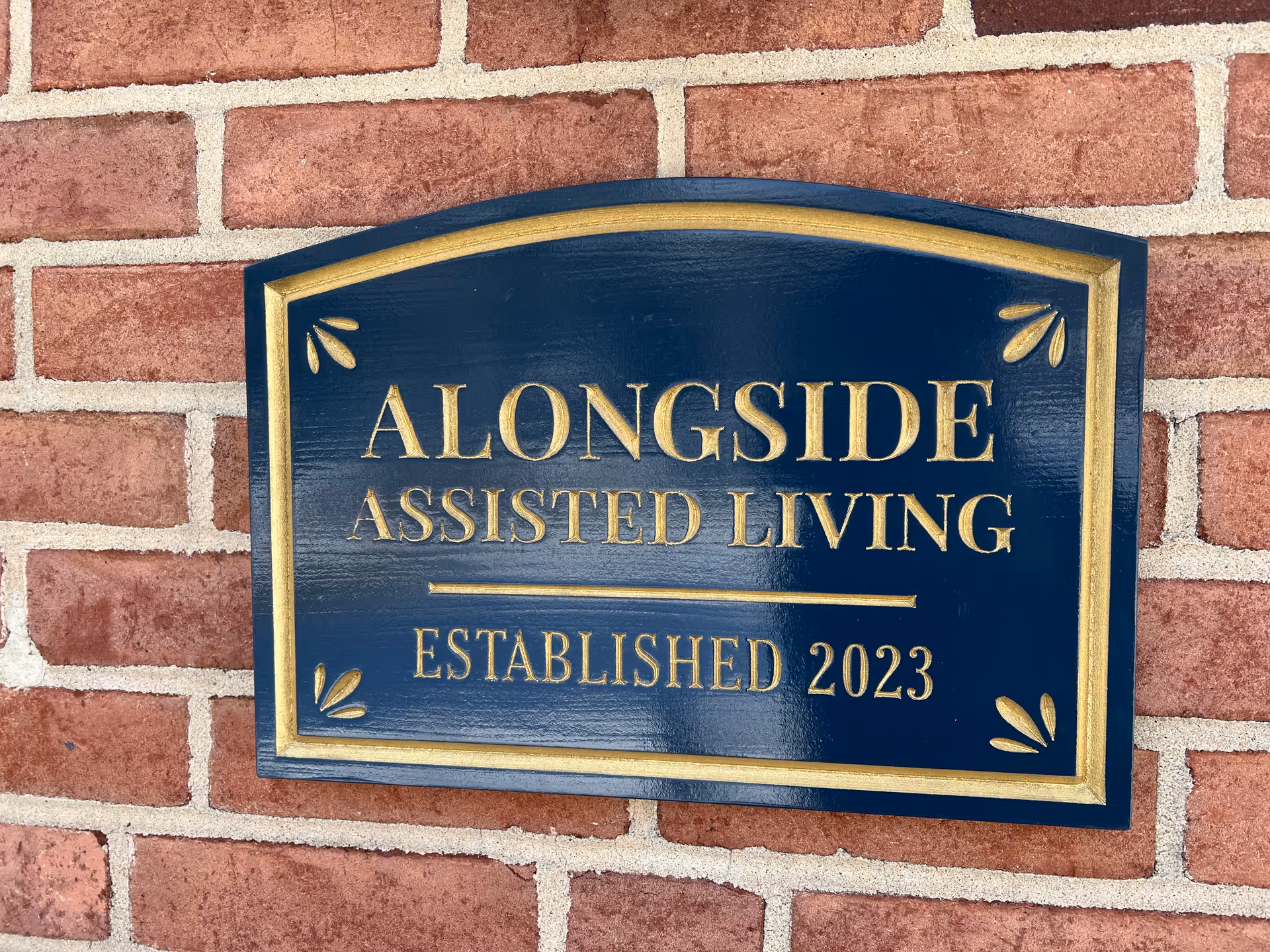 A blue and gold sign mounted on a red brick wall that reads 'ALONGSIDE ASSISTED LIVING ESTABLISHED 2023'.