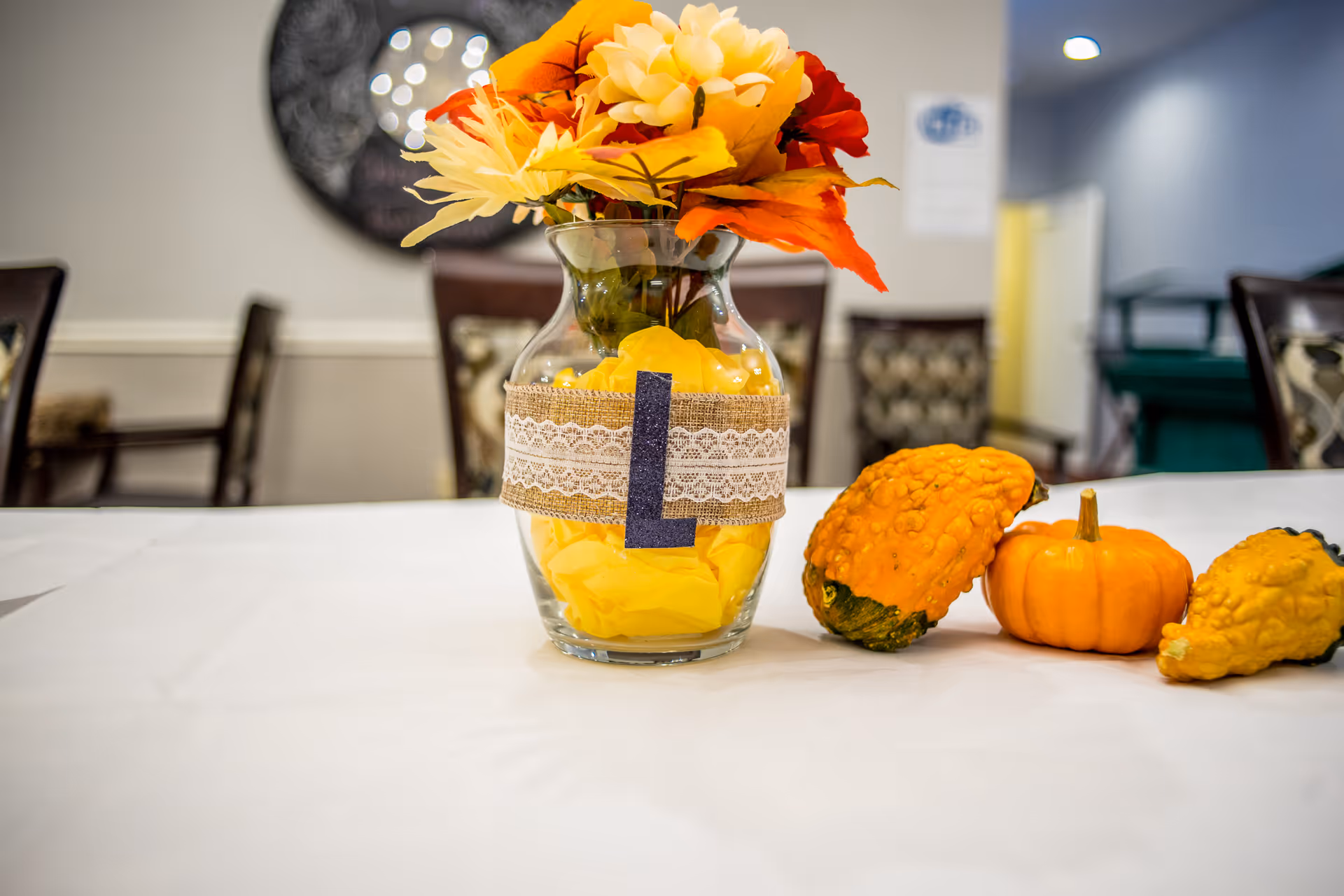 A glass vase decorated with a burlap and lace ribbon and a large letter L, filled with yellow tissue paper and artificial flowers in yellow, orange, and red colors, placed on a white tablecloth. Next to the vase are three small decorative gourds and a pumpkin. The background shows blurred chairs and a wall with a circular decorative mirror.