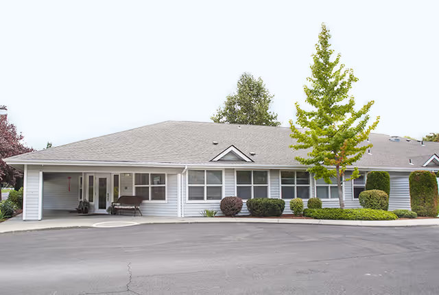 Single-story building with a gray roof and white siding, surrounded by neatly trimmed bushes and a small tree in front. The building has multiple windows and a covered entrance with a bench nearby. The area in front is paved with asphalt.
