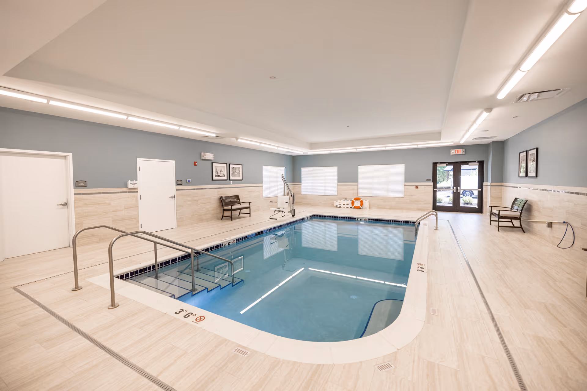 Indoor swimming pool area with light-colored tile flooring and walls, blue water, metal handrails, pool lift, benches along the walls, and large windows with blinds. The room has a high ceiling with recessed lighting and an exit door at the far end.