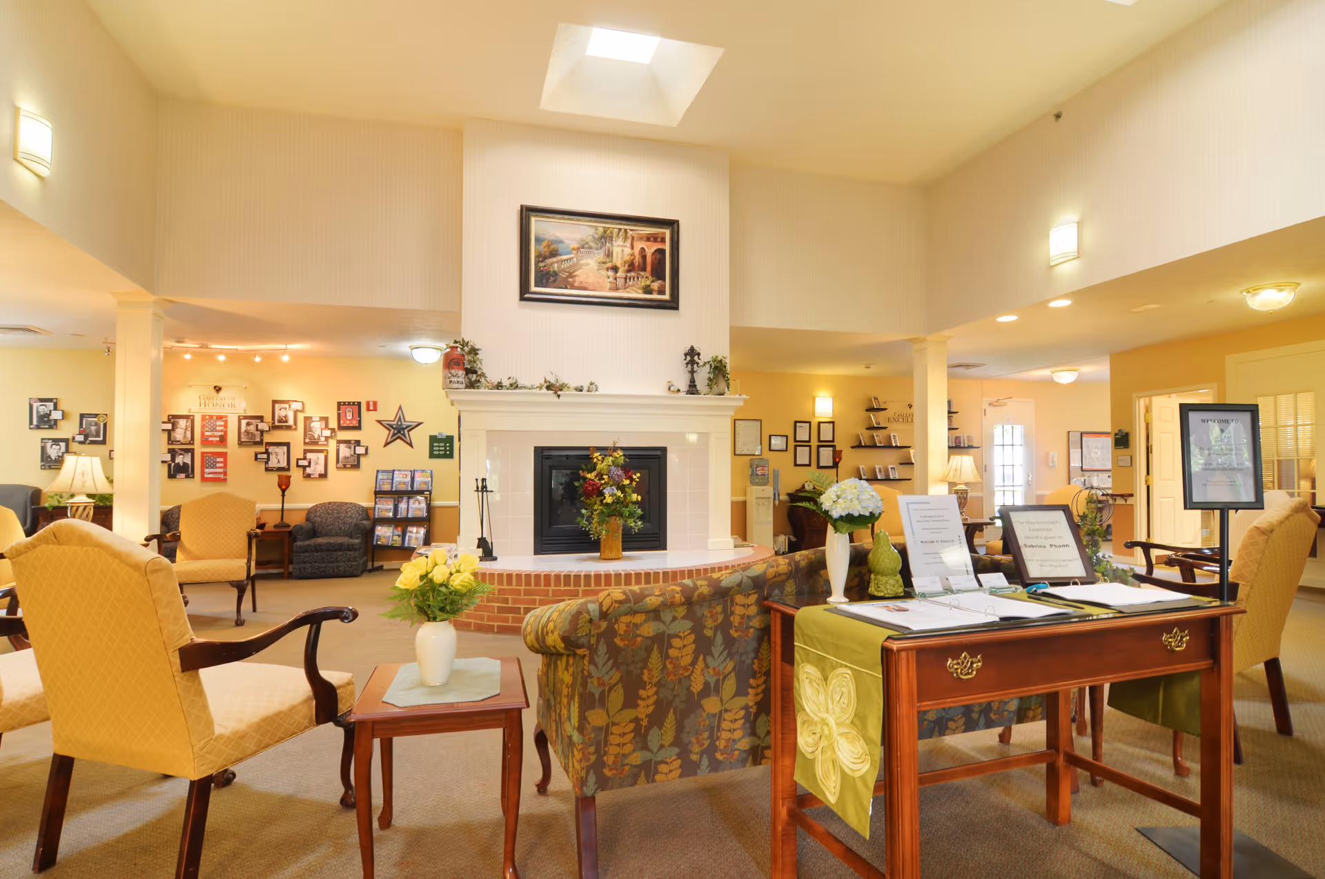 A cozy senior living facility common area with a central fireplace, surrounded by comfortable armchairs and a floral-patterned sofa. The room is warmly lit with wall sconces and ceiling lights, decorated with framed pictures, plants, and a painting above the fireplace. A wooden table with informational materials and flowers is positioned in the foreground.