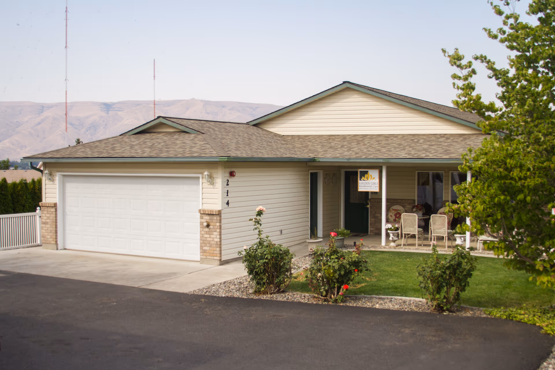 Exterior view of a single-story residential building with a two-car garage, a small front porch with chairs, a green lawn, and some bushes and trees. Mountains are visible in the background under a clear sky.