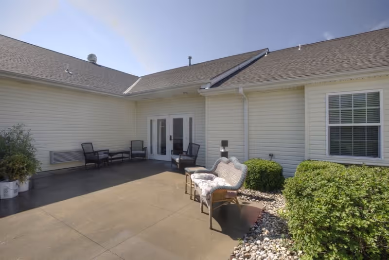 Outdoor patio area at a senior living facility with several chairs and a cushioned bench arranged on a concrete surface. The building has light-colored siding, a window with blinds, and a set of double glass doors. There are green bushes and decorative rocks along the edge of the patio under a clear blue sky.