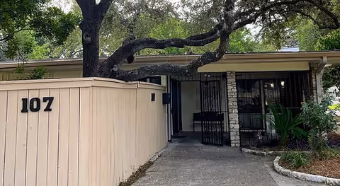 Exterior view of a senior living facility entrance with a beige wooden fence displaying the number 107, a large tree with sprawling branches, and a stone and metal gate leading to the building.