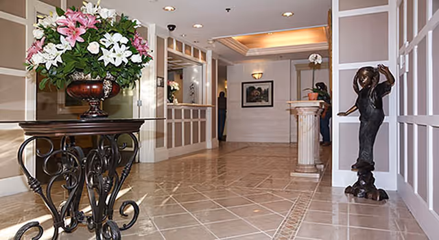 A well-lit interior hallway with tiled floors and decorative wall paneling. On the left, there is a round table with a large floral arrangement of pink and white lilies. On the right, there is a bronze statue of a person wearing a hat. The hallway leads to a reception area with a counter and a framed picture on the far wall.
