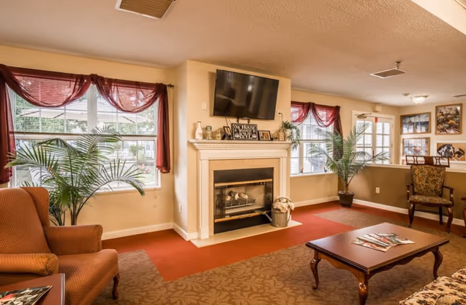 Cozy senior living room lounge with a fireplace, wall-mounted TV, armchairs, coffee table, and large windows with red curtains.