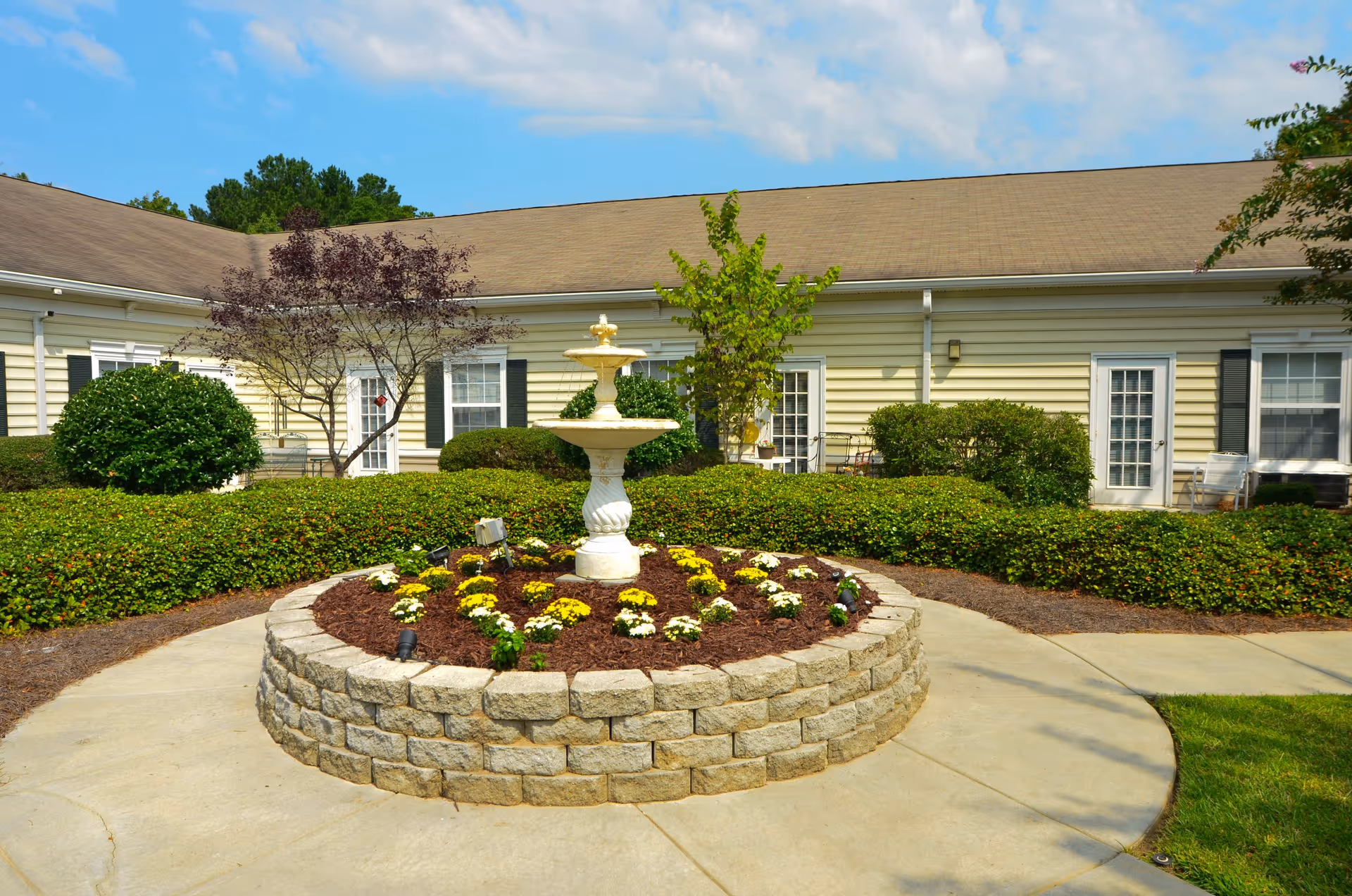 Outdoor courtyard area at Lancaster Grove Senior Living featuring a circular raised flower bed with a white tiered fountain in the center, surrounded by neatly trimmed bushes and small trees, with a beige building and multiple doors and windows in the background under a blue sky.