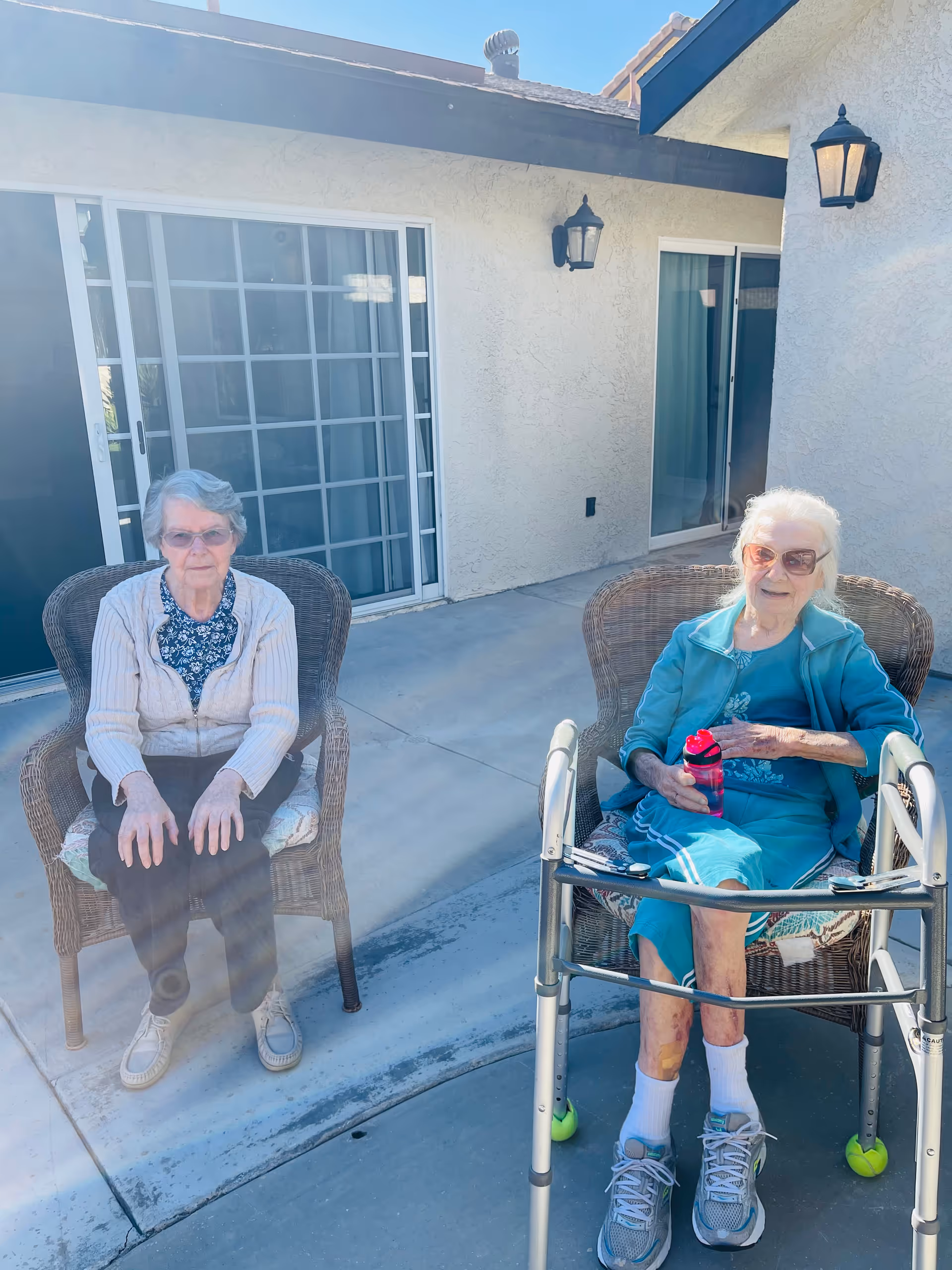 Two elderly women seated on wicker chairs on a sunny patio outside a residential building, one using a walker and holding a water bottle.