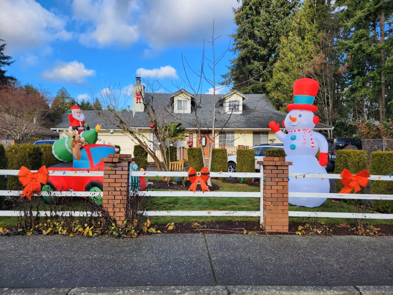 Front yard of a house decorated with Christmas inflatables including a snowman wearing a red and green hat and a red truck carrying Santa Claus and a reindeer. The yard is enclosed by a white fence with brick pillars, adorned with red bows and string lights. The house has a gray roof with two dormer windows and a chimney with a red 'W' decoration. Trees and bushes surround the property under a partly cloudy blue sky.
