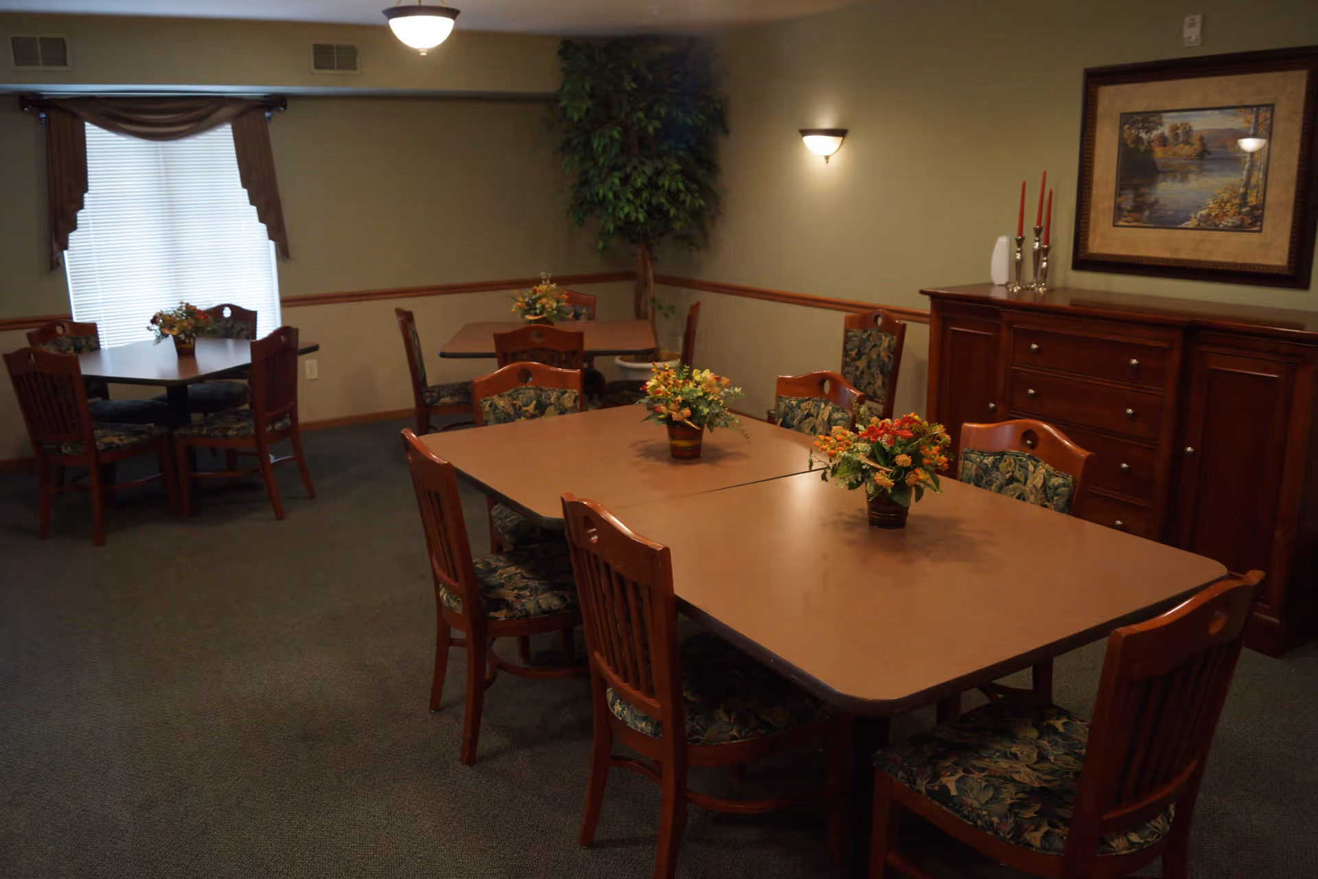 Interior view of a dining room with multiple tables and wooden chairs with floral cushions. Each table has a small floral centerpiece. The room has beige walls with a wooden trim, a window with blinds and a valance, a wooden sideboard with decorative items, a framed painting on the wall, and a potted plant in the corner.
