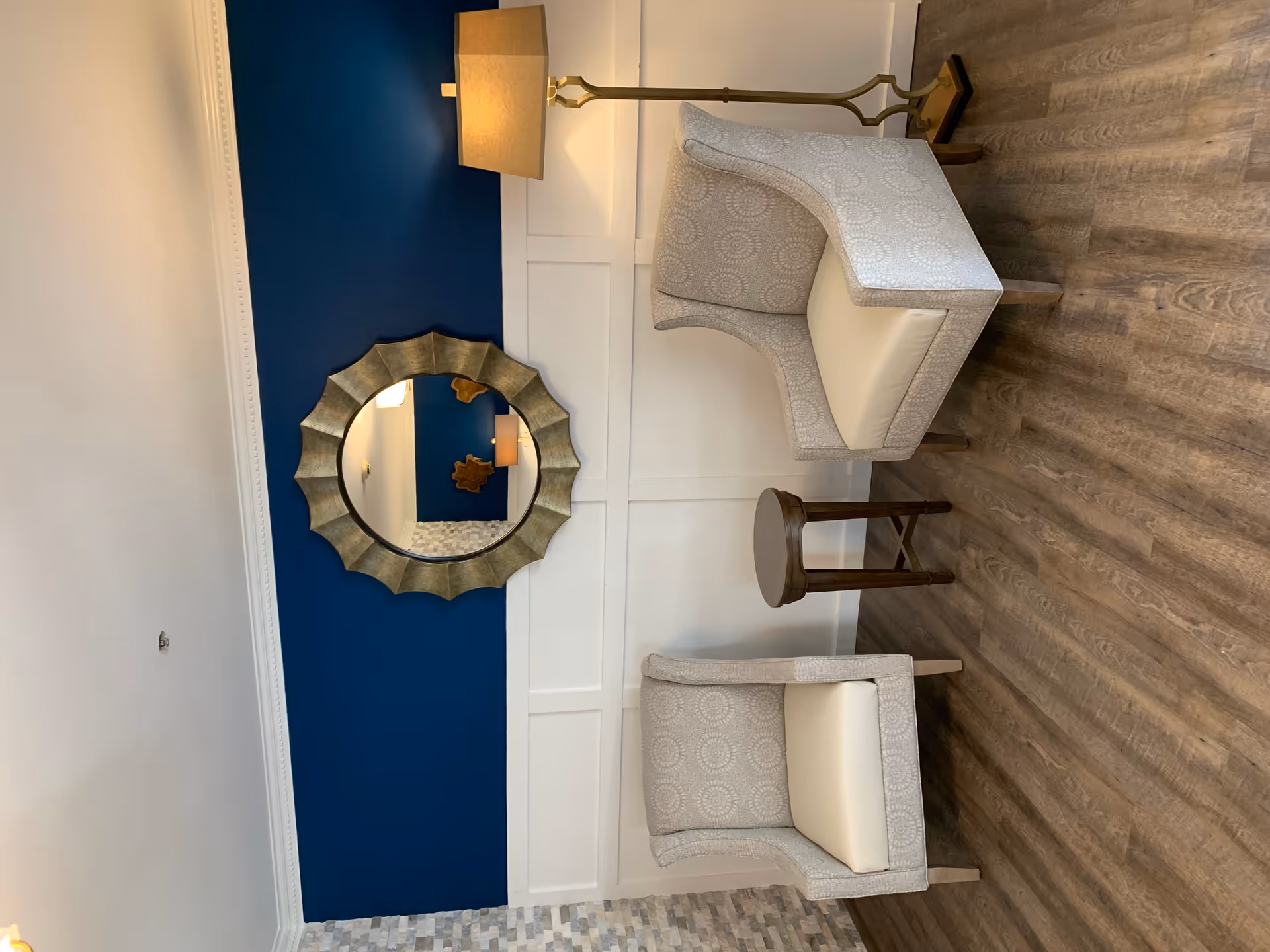 A cozy seating area with two patterned armchairs and a small round wooden table between them. A floor lamp with a beige shade stands behind one chair. The wall behind features a decorative round mirror with a gold frame, set against a blue and white paneled wall. The floor is covered with wood-style flooring.